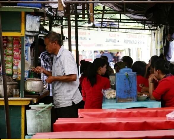 Warung makan sederhana di pinggir jalan yang ramai dikunjungi wisatawan, dengan penjual sedang melayani pelanggan.